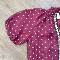 A close-up of a burgundy top with white polka dots, featuring a smocked elasticated yoke and a patterned side panel, laid flat on a gray wood floor.