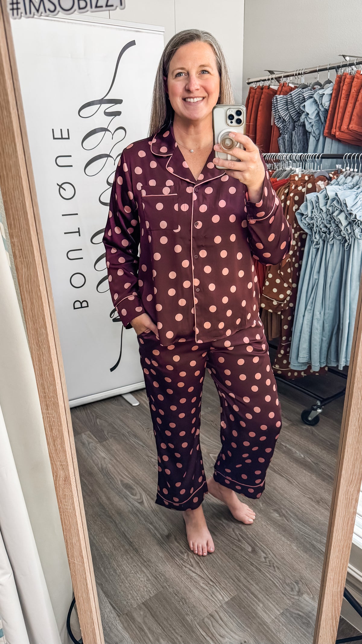 A woman takes a mirror selfie wearing a matching two-piece pajama set with a burgundy base and pink polka dots. The setting is a boutique clothing area with a clothing rack and a white sign in the background.