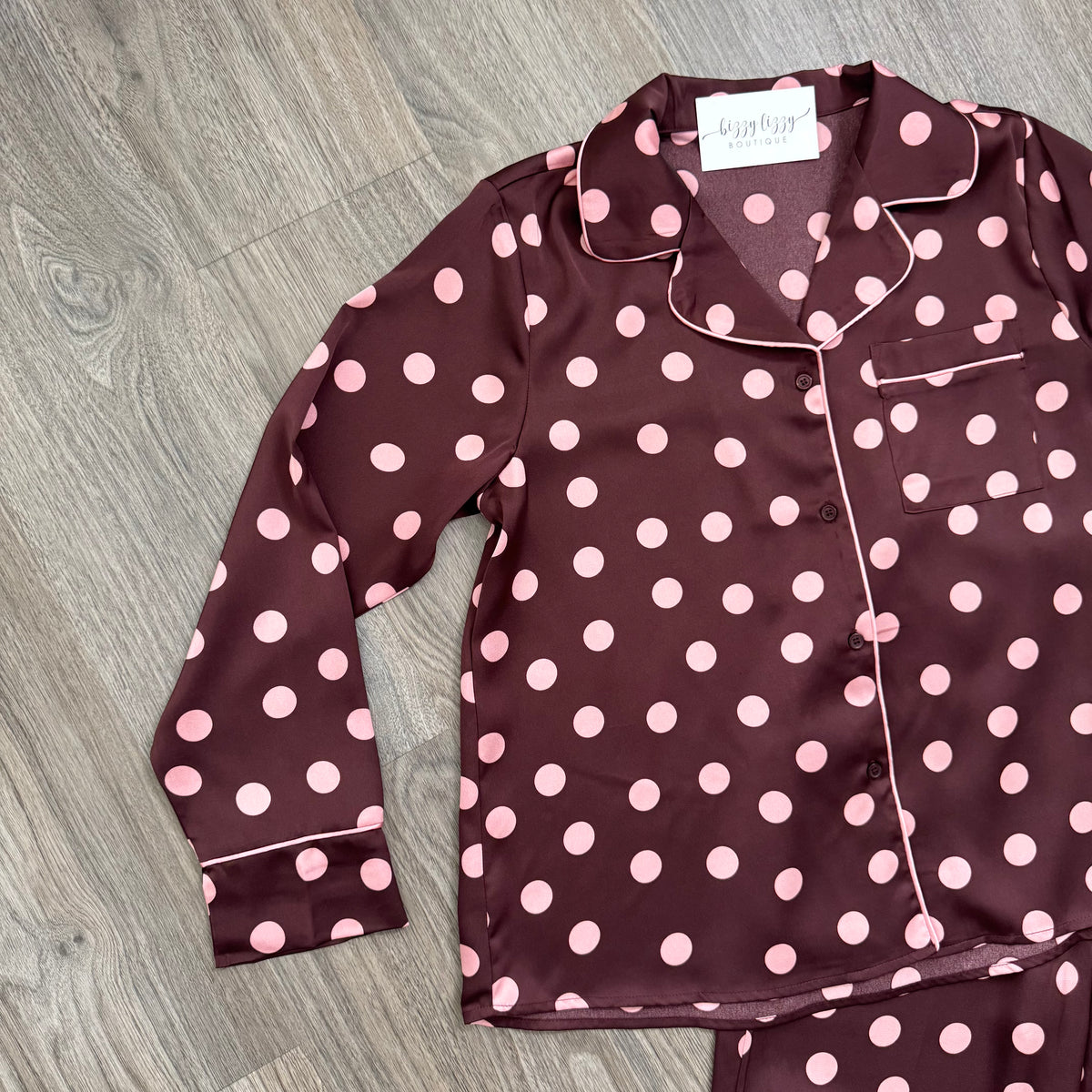 A long-sleeve pajama top laid flat on a gray wood-look floor, featuring a dark burgundy base with pink polka dots and light piping. A matching pair of pajama bottoms is partially visible at the bottom right.