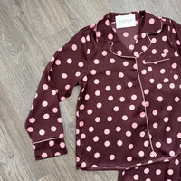 A long-sleeve pajama top laid flat on a gray wood-look floor, featuring a dark burgundy base with pink polka dots and light piping. A matching pair of pajama bottoms is partially visible at the bottom right.