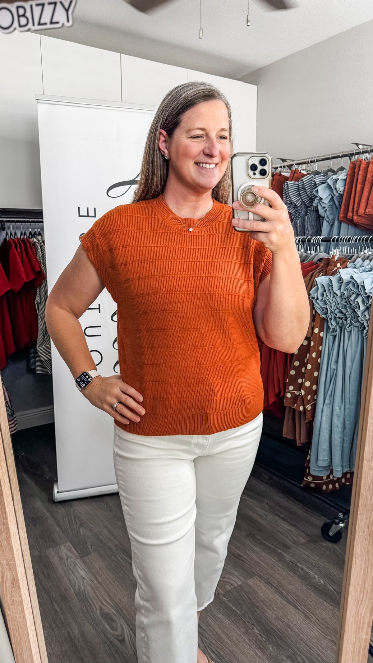 A woman takes a mirror selfie in a boutique wearing a textured orange short-sleeve knit top and white pants. The boutique interior has clothing racks and a white display board in the background.