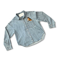 A light blue denim long-sleeve button-up shirt with a small embroidered horse head in the chest pocket, displayed flat on a white background.