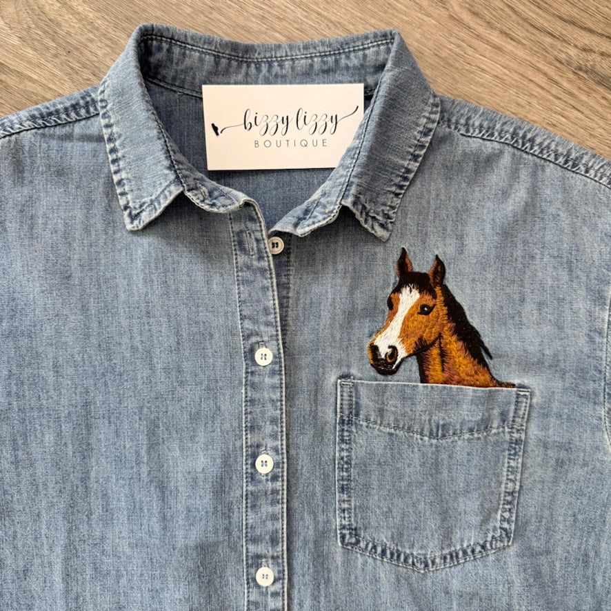 A light blue denim button-up shirt laid flat on a wood surface, featuring a printed embroidered horse head on the left chest pocket. The inside collar shows a brand label reading 'bizzy lizzy BOUTIQUE'.