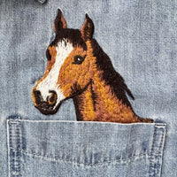 Close-up of a brown embroidered horse head patch with a white blaze, placed on light blue denim fabric above a front pocket.