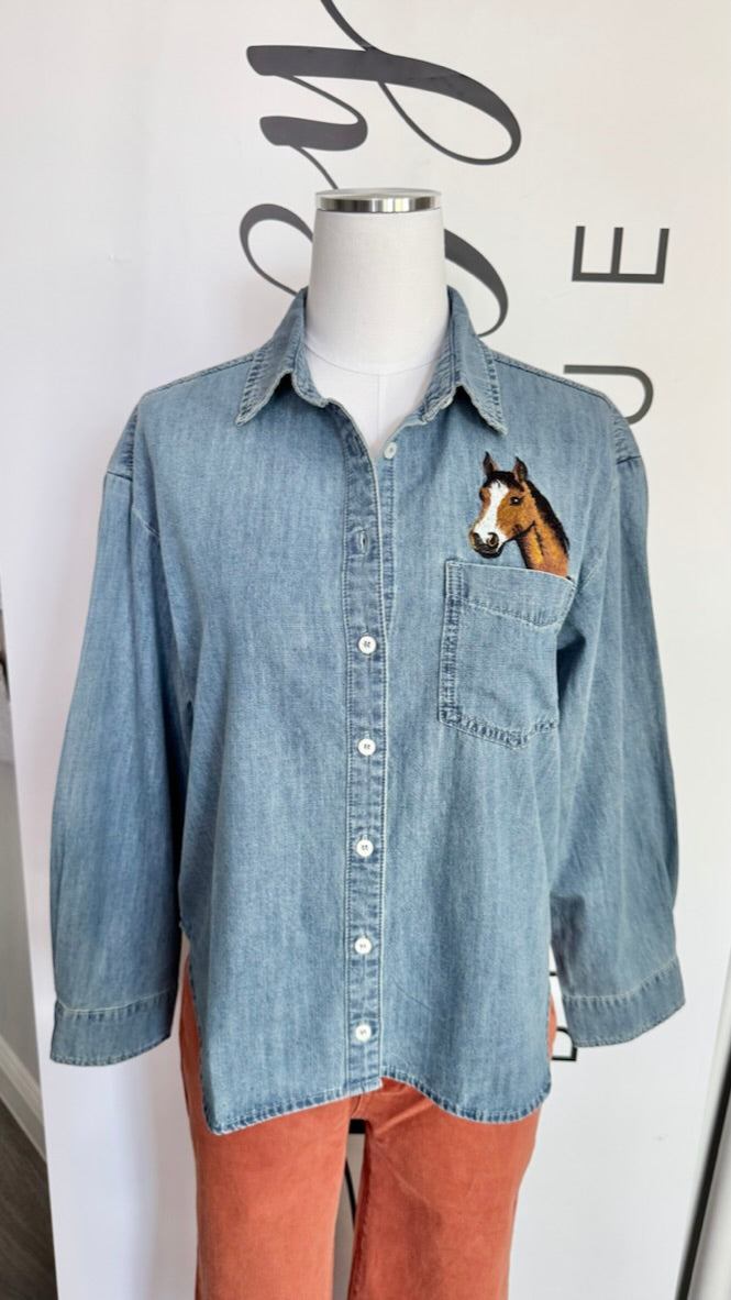 A light blue denim button-up shirt displayed on a mannequin torso, featuring a printed horse face graphic on the left chest pocket and orange pants underneath.