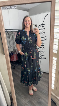 A woman takes a mirror selfie in a boutique showing a black midi dress with colorful floral embroidery. The boutique interior has gray wood floors, a white wall with black script, and a clothing rack in the background.