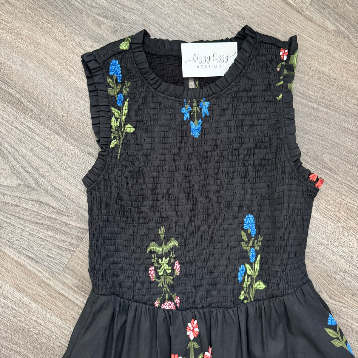 A sleeveless black smocked dress with ruffled trim and colorful embroidered wildflower motifs, laid flat on a gray wood-look floor. A white brand label reading 'buzzy bee boutique' is visible at the neckline.