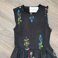 A sleeveless black smocked dress with ruffled trim and colorful embroidered wildflower motifs, laid flat on a gray wood-look floor. A white brand label reading 'buzzy bee boutique' is visible at the neckline.