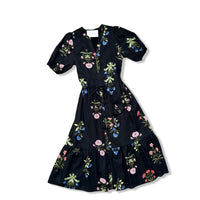 A black midi dress with puff sleeves, a V-neckline, and a tiered ruffle hem, decorated with a colorful floral and botanical print in pink, blue, green, and yellow.