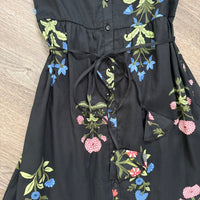 A black sleeveless dress with a floral print featuring blue, pink, and green flowers, laid flat on a light wood floor showcasing the decorative tie belt.