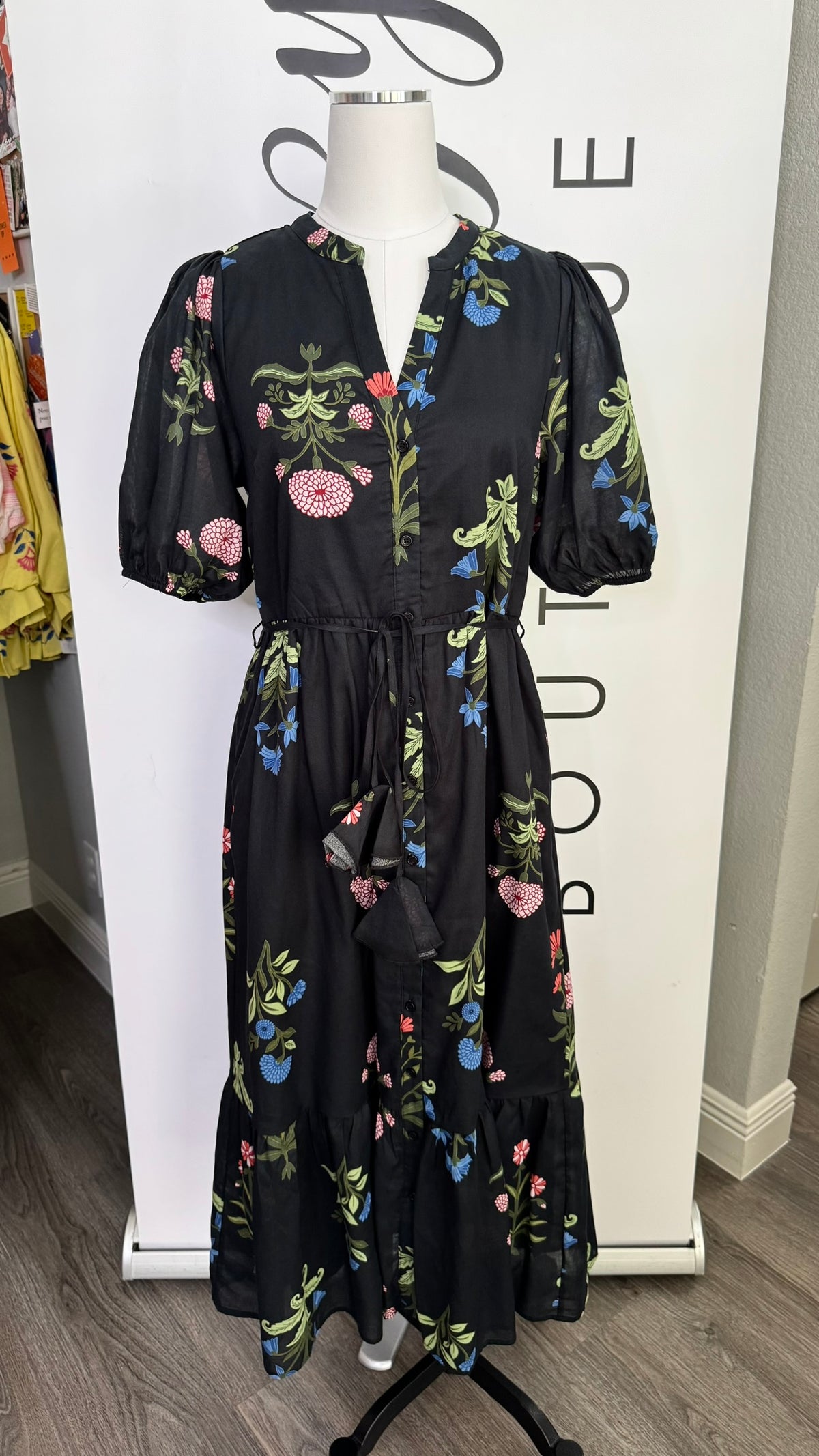 A black midi dress with a colorful floral print, displayed on a mannequin in front of a white boutique sign.