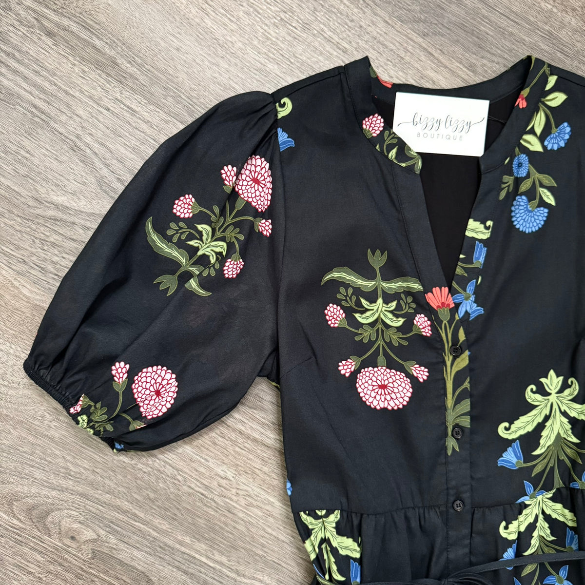 A black short-sleeve blouse with puff sleeves and a colorful floral print, laid flat on a gray wood-textured surface. The print features pink, blue, and green flowers and leaves.