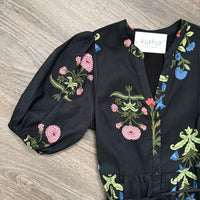 A black short-sleeve blouse with puff sleeves and a colorful floral print, laid flat on a gray wood-textured surface. The print features pink, blue, and green flowers and leaves.