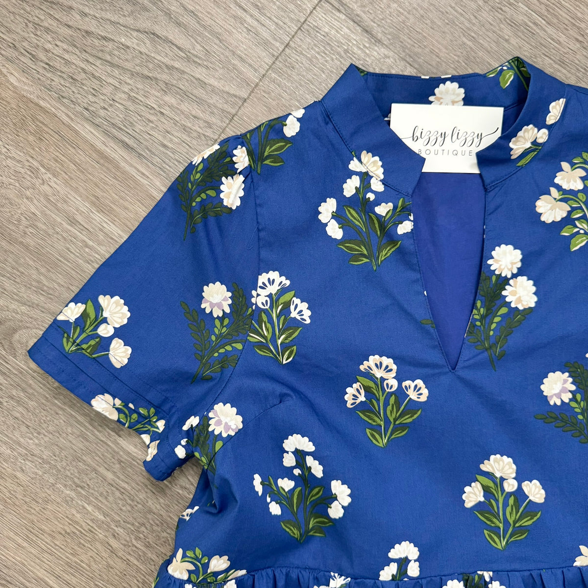 A royal blue short-sleeve dress with a white and green floral print, laid flat on a gray wood-look floor. The top features a split V-neckline with a white paper brand label inside that reads 'Bizzy Lizzy Boutique'.