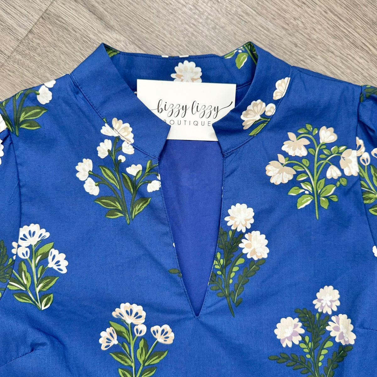 A royal blue dress with a white floral print and green stems, featuring a split V-neckline. A white brand label reading 'bizzy lizzy BOUTIQUE' is visible at the collar, and the garment is laid flat on a gray wood-textured surface.