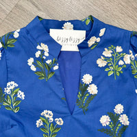A royal blue dress with a white floral print and green stems, featuring a split V-neckline. A white brand label reading 'bizzy lizzy BOUTIQUE' is visible at the collar, and the garment is laid flat on a gray wood-textured surface.
