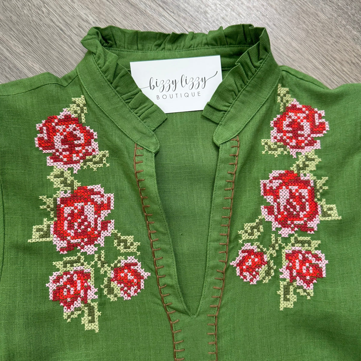 A close-up of a green blouse with red and pink embroidered roses and green leaves on the front, featuring a split neckline and a white neck label that reads 'bizzy lizzy boutique'.