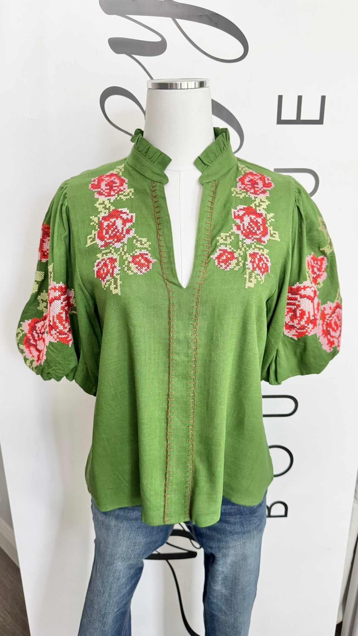 A green short-sleeve blouse with red and white floral embroidery on the chest and sleeves, displayed on a mannequin torso with blue jeans against a white background with black lettering.