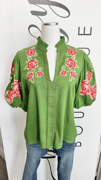 A green short-sleeve blouse with red and white floral embroidery on the chest and sleeves, displayed on a mannequin torso with blue jeans against a white background with black lettering.