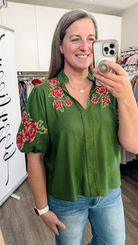 A woman takes a mirror selfie in a boutique while wearing a green short-sleeve button-up blouse with red floral embroidery on the chest and sleeve, paired with blue jeans.
