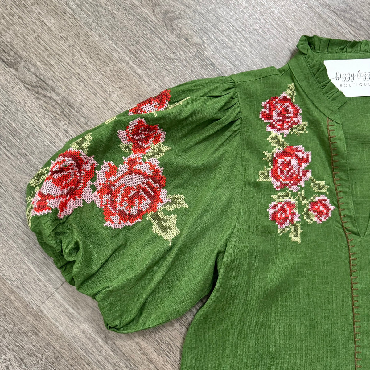A green blouse with puff sleeves and embroidered red roses on the sleeve and chest, laid flat on a gray wood-textured surface. A white neck label reads 'bizzy lizzyy'.