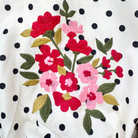 A close-up of a white fabric with black polka dots, featuring a prominent embroidered floral motif with red and pink flowers, green leaves, and mustard-yellow accents.