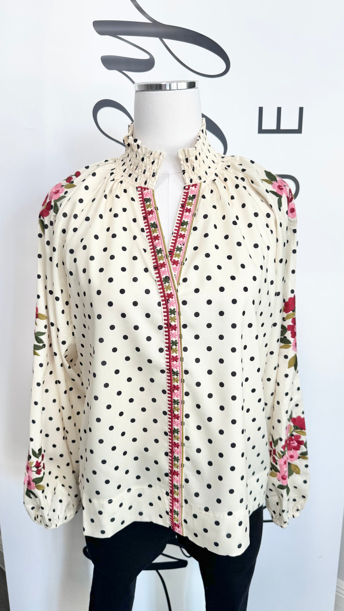 A cream-colored long-sleeve blouse with black polka dots, featuring a pink and red embroidered front placket and floral patterns on the shoulders and sleeves, displayed on a mannequin torso.