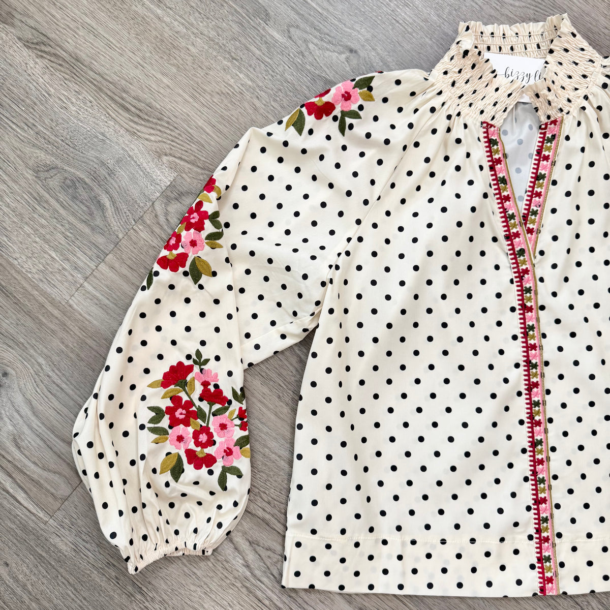 An off-white blouse with black polka dots, featuring red and pink floral patterns on the sleeves and a decorative red and green embroidered trim along the front zipper.