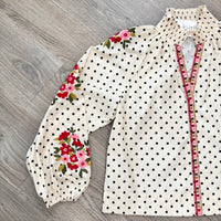 An off-white blouse with black polka dots, featuring red and pink floral patterns on the sleeves and a decorative red and green embroidered trim along the front zipper.