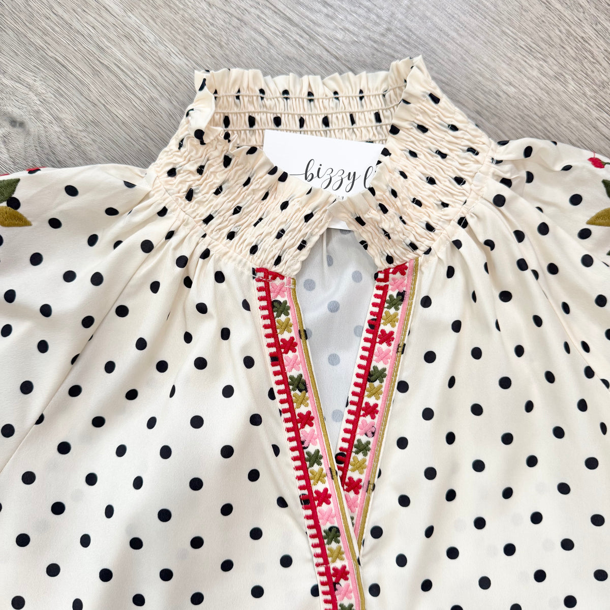 An off-white blouse with black polka dots, featuring a smocked high collar and a V-neckline trimmed with pink, green, and red floral embroidery.