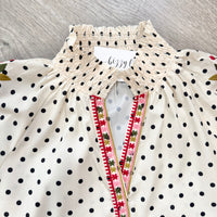An off-white blouse with black polka dots, featuring a smocked high collar and a V-neckline trimmed with pink, green, and red floral embroidery.
