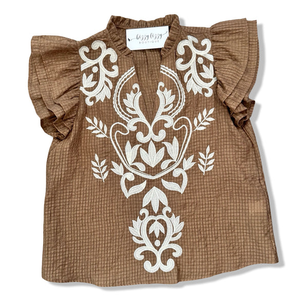 A brown textured cotton blouse with white embroidered floral patterns on the front, featuring ruffled short sleeves and a V-neckline, laid flat on a white background.