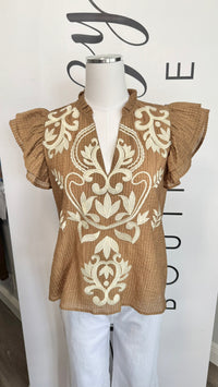 A brown short-sleeve blouse with cream damask embroidery and ruffled sleeves, displayed on a mannequin torso with white pants against a white boutique backdrop.