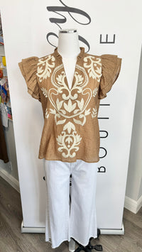 A tan short-sleeve blouse with cream damask embroidery and ruffled sleeves, displayed on a mannequin with white pants in front of a boutique backdrop.