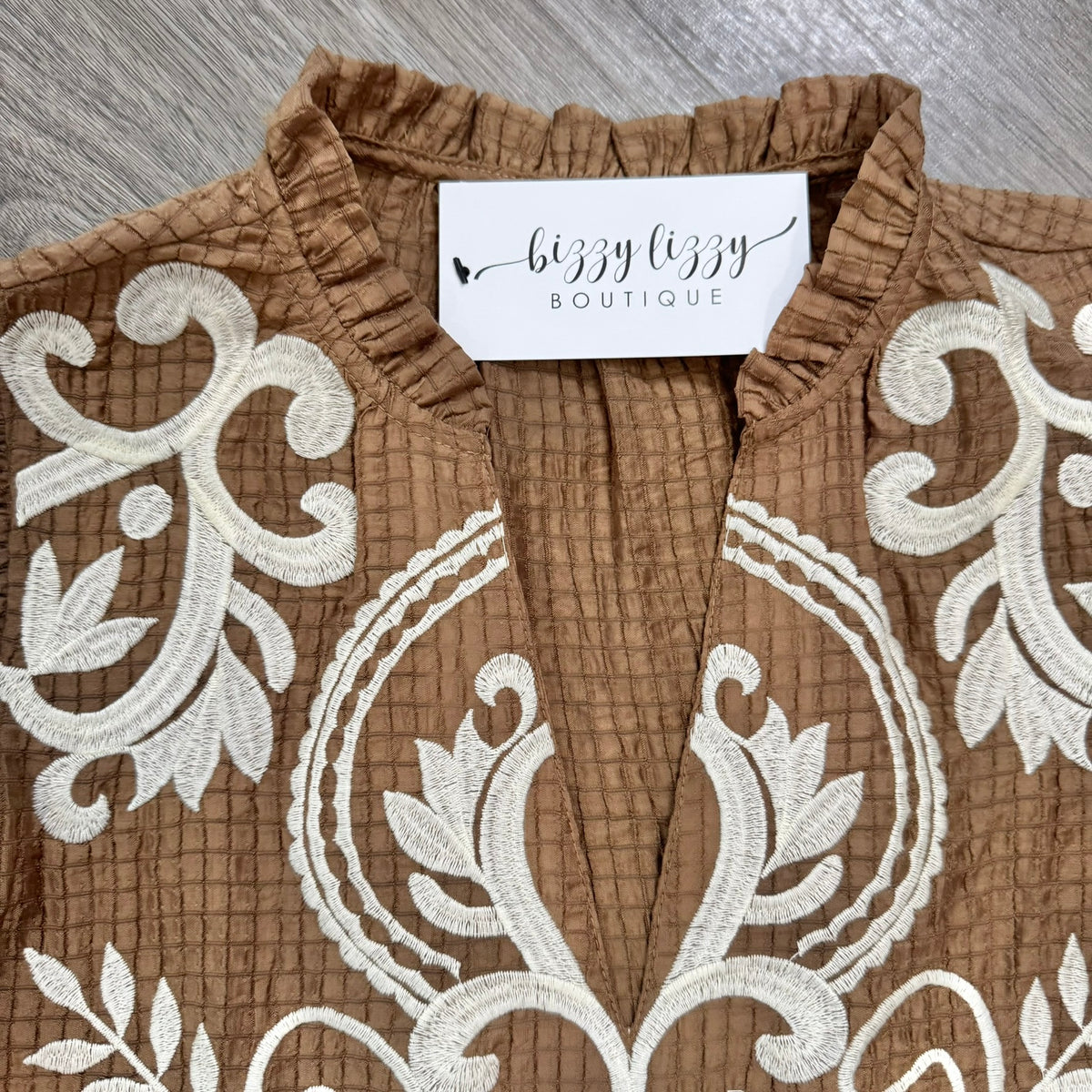 A close-up of a brown textured top with white embroidered scrollwork, featuring a ruffled collar and a visible 'bizzy lizzy BOUTIQUE' label.