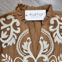 A close-up of a brown textured top with white embroidered scrollwork, featuring a ruffled collar and a visible 'bizzy lizzy BOUTIQUE' label.