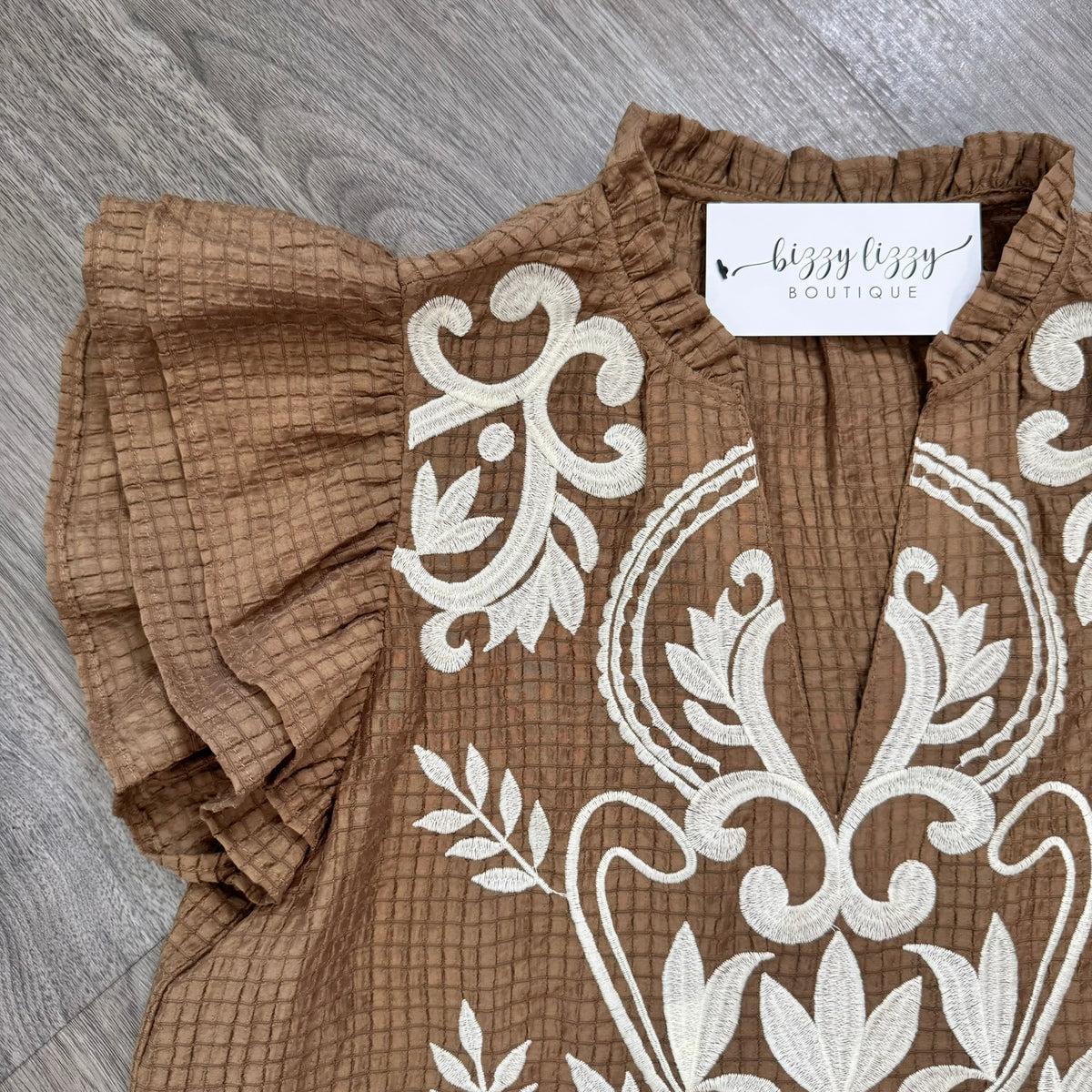 A close-up of a brown textured blouse with white floral embroidery and ruffled sleeves, laid flat on a gray wood floor. A white neck label reads 'bizzy lizzy BOUTIQUE'.
