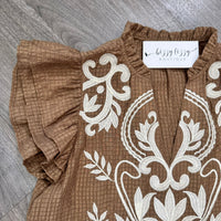 A close-up of a brown textured blouse with white floral embroidery and ruffled sleeves, laid flat on a gray wood floor. A white neck label reads 'bizzy lizzy BOUTIQUE'.