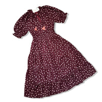 A dark burgundy midi dress with pink polka dots, featuring short puff sleeves, a ruffled neckline with a tie, and a smocked waist. The dress is displayed flat against a white background.
