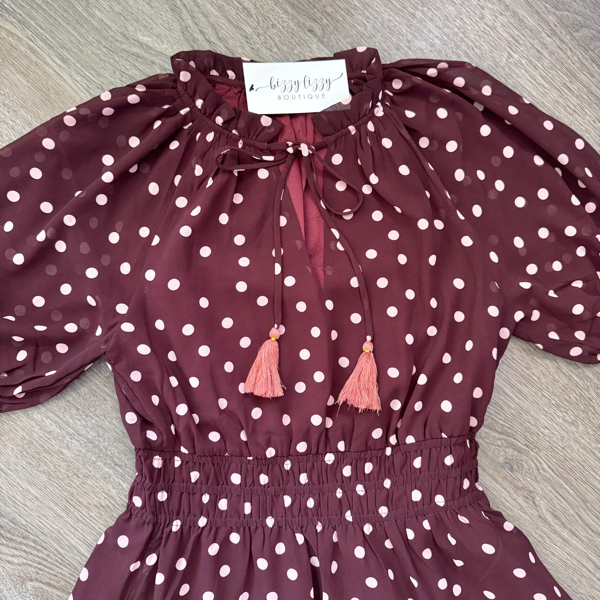 A burgundy and pink polka dot dress with puff sleeves, a tie-neck bow, and pink tassel accents, laid flat on a gray wood-textured background. A brand tag reading 'buzzy bee boutique' is visible inside the collar.