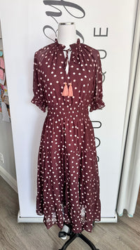 A dark burgundy midi dress with pink polka dots, featuring a high neck with a tie-front closure and two pink tassel accents, displayed on a mannequin in front of a white branded backdrop.