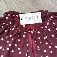 A close-up of a burgundy blouse with pink polka dots, featuring a ruffled neckline, a front tie closure, and a white paper label that reads 'bizzy lizzy BOUTIQUE'.