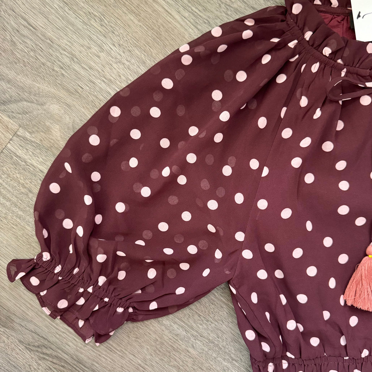 A close-up of a child's burgundy puff-sleeve blouse with pink  polka dots, featuring elasticized cuffs and a drawstring waist, laid flat on a gray wood floor.