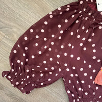 A close-up of a child's burgundy puff-sleeve blouse with pink  polka dots, featuring elasticized cuffs and a drawstring waist, laid flat on a gray wood floor.