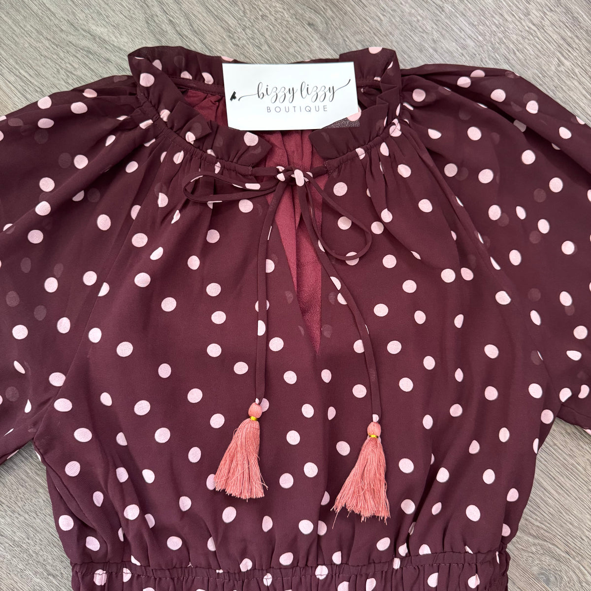 A burgundy long-sleeve blouse with pink  polka dots, featuring a tie-neck design with pink tassels and a white neck label that reads 'Bijoux Boutique'.