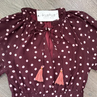 A burgundy long-sleeve blouse with pink  polka dots, featuring a tie-neck design with pink tassels and a white neck label that reads 'Bijoux Boutique'.