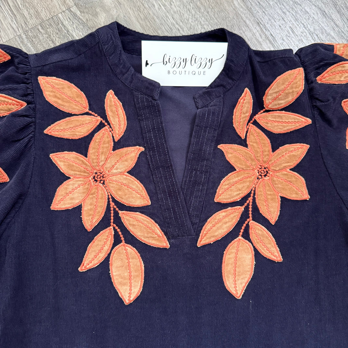 A close-up of a navy blue corduroy top with orange embroidered floral applique patterns on the chest, featuring a split neckline and puff sleeves. A white neck label reads 'bizzy lizzy BOUTIQUE'.