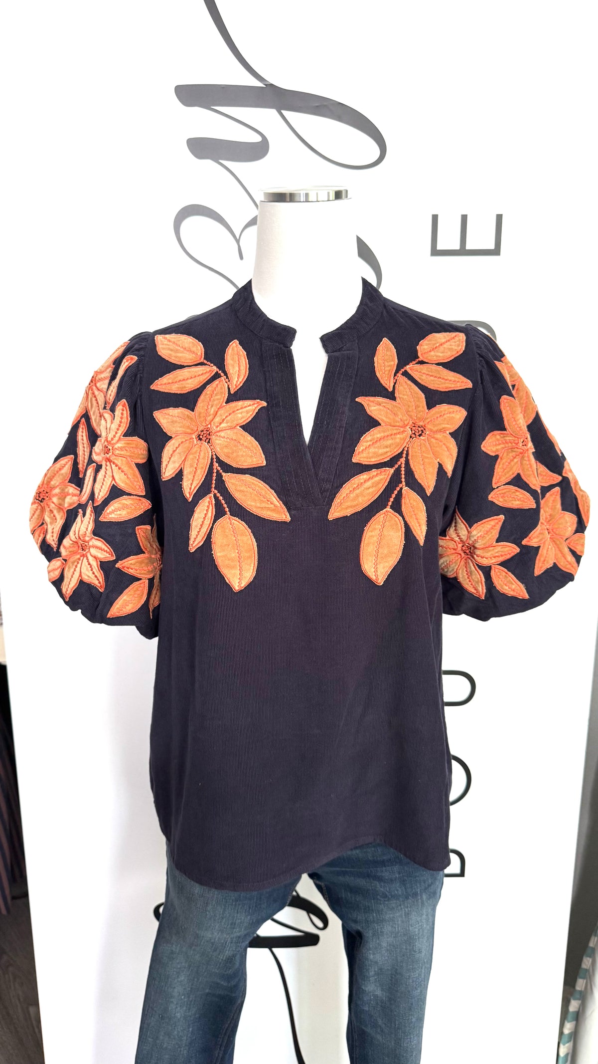 A dark navy blue short-sleeve top with orange floral embroidery on the bodice and sleeves, displayed on a mannequin torso with blue jeans against a white background.