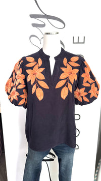 A dark navy blue short-sleeve top with orange floral embroidery on the bodice and sleeves, displayed on a mannequin torso with blue jeans against a white background.
