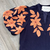 A navy blue blouse with orange embroidered floral applique patterns on the sleeves and chest, laid flat on a gray wood-textured surface. A white neck label reads 'bizzy lizzy BOUTIQUE'.
